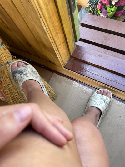 The image shows a person's legs and feet wearing cartoon-themed slides. The person is sitting on the ground in front of a wooden doorway and a wooden deck. To the left of the person is a broom made of straw with a blue tie. The ground is covered in cardboard. The environment suggests an outdoor setting, possibly a backyard or porch area. The user is wearing a floral print shirt. The location is identified as Сады Им. Володарского Снт, Russia.