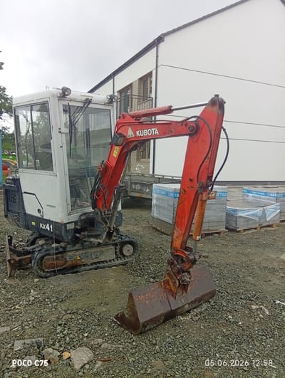 A Kubota KX41 mini excavator, featuring a white cabin and a red boom arm with a rusty bucket, is the central subject of the image. The excavator's cabin has glass windows, a white roof with two lights, and windshield wipers. The black rubber tracks are visible, and a dozer blade is attached to the front base. Visible text on the excavator includes "KUBOTA" on the boom arm, "KX41" on the side of the engine compartment, and "H. HEIMBUCHER" on the cabin roof.

The excavator is positioned on a ground covered with gravel and small rocks, suggesting a construction or development site. In the background, to the right, is a modern, two-story white building with a dark grey roofline and some visible windows, possibly indicating a residential or commercial structure. Several pallets stacked with grey paving stones or tiles, wrapped in clear plastic with blue bands displaying "BETARD" and "RETARD" text, are located between the excavator and the building. A small utility trailer is partially visible behind the excavator's boom.

The sky is overcast, indicating a cloudy daytime setting. No people are visible in the scene. The overall setting is an outdoor worksite in Kiełczów, Poland. The image has a timestamp of "05.06.2026 12:58" and a device watermark "POCO C75" at the bottom.
