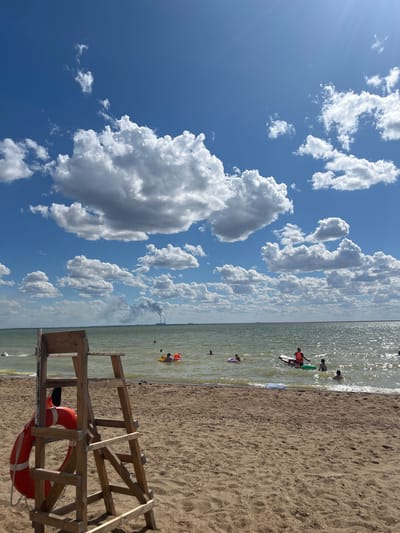 In a sunlit outdoor scene at a beach in Gres-2, Kazakhstan, a wooden lifeguard chair stands prominently on the sandy shore, adorned with a bright orange life preserver. Beyond the chair, the calm, shimmering waters of a large lake or sea stretch towards the horizon under a vivid blue sky populated with fluffy white clouds. Several people are visible in the water, some swimming, others floating on inflatable rings or boards. In the distance, industrial towers with plumes of smoke rise against the skyline, indicating the presence of a power plant or industrial facility. The overall atmosphere is one of a leisurely summer day at the beach.