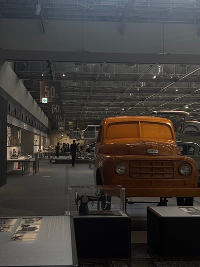 The image depicts the interior of the Toyota Commemorative Museum of Industry and Technology in Nagoya, Japan, with a focus on the automotive exhibits. In the foreground, a large, orange wooden model of a truck, likely a historical Toyota model, dominates the right side of the frame. To the left of the truck, a sewing machine is displayed under a glass case on a black pedestal. In the background, other vehicle chassis and historical exhibits are visible, alongside a group of visitors observing the displays. Overhead, the museum's industrial ceiling structure with exposed beams and lighting is apparent. Signs indicating "50s" and "60s" hang from the ceiling, marking different exhibit sections. The environment is well-lit, showcasing the various artifacts and the architectural features of the museum.
