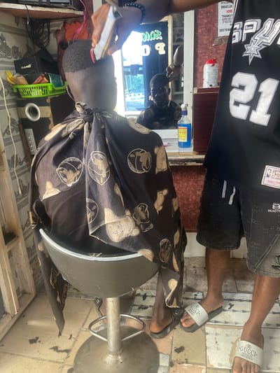In Kporva, Nigeria, a barber is trimming the hair of a young boy in a salon. The boy sits in a chair covered by a cape adorned with a teddy bear pattern. The barber, wearing a black basketball jersey and denim shorts, uses clippers to cut the boy's hair. The salon appears to be a small, modestly equipped space with shelves displaying various hair products and tools. A mirror reflects another person in the background. The floor is tiled, and the overall atmosphere suggests a local, community-based barbershop.