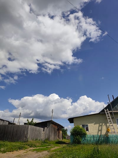 The image captures a sunny day in the village of Kvatsa, Russia. The foreground features a dirt path bordered by lush green grass and vegetation. To the left, a weathered wooden fence encloses a property with a dark wooden shed visible behind it. In the center and right of the frame, a light-colored house with a green trim fence stands prominently. A tall wooden ladder leans against the house, and a yellow gas meter is mounted on the wall. A dense green bush grows near the house. The background is dominated by a vibrant blue sky filled with large, fluffy white clouds. Electrical wires and utility poles are also visible, crisscrossing the sky. The overall scene depicts a typical rural residential setting.