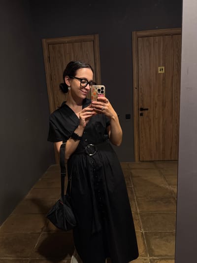 A woman with dark hair pulled back and wearing glasses is taking a mirror selfie. She is wearing a black collared dress with buttons down the front and a black belt with a silver buckle. She has a black smartwatch on her left wrist and is carrying a black bag with a strap over her shoulder. The background appears to be a hallway with two wooden doors. One door on the right has a gender-neutral restroom sign. The floor is tiled. This image was taken in Bakhtiyary, Russia.