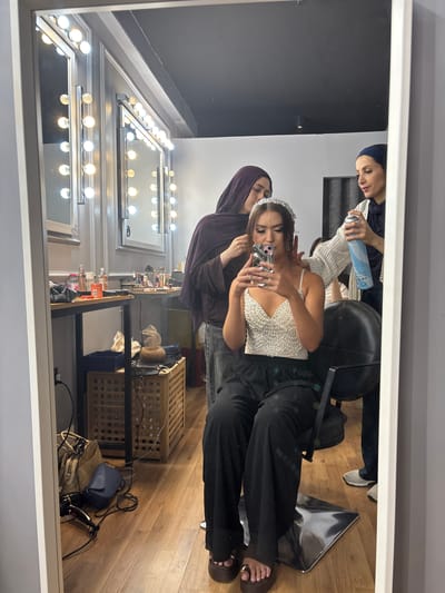 The image captures a reflection in a mirror of a beauty salon environment, likely located in Sweileh, Jordan. In the foreground of the reflection, a young woman sits in a black salon chair, holding a smartphone and taking a selfie. She wears a sparkly white top, black pants, and platform sandals. A tiara is positioned on her head, suggesting preparation for a special event. Two other individuals, presumably makeup artists or stylists, are attending to her. One, wearing a purple headscarf, appears to be adjusting her hair. The other, in a dark headscarf and striped shirt, is holding a can of hairspray. The background of the salon features a vanity mirror with numerous lights, makeup products on a table, and a woven laundry basket containing items. The floor is wooden. The overall scene depicts a moment of preparation and styling.