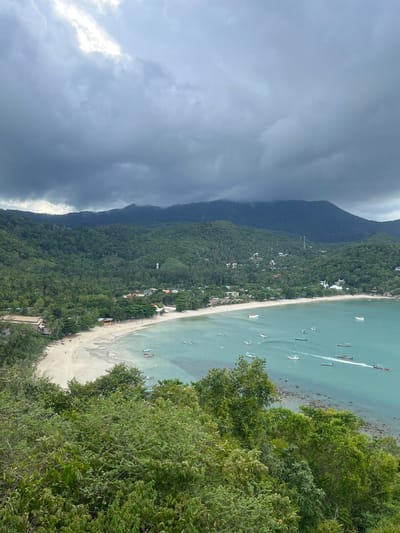 An elevated, wide-angle view captures a coastal bay and an adjacent settlement under an overcast sky in Tambon Ban Tai, Thailand. The upper third of the image is dominated by heavy, dark grey cloud cover, with a brighter, less dense area visible on the far left. Below the clouds, a range of verdant, densely forested mountains and hills extends across the horizon, with some peaks partially shrouded in mist or lower clouds.
The midground features a crescent-shaped bay with clear, turquoise-to-light-blue water. A light sandy beach borders the bay's inner curve, where a small coastal village is situated. The village comprises numerous buildings, including residential structures, small commercial establishments, and a distinct white-domed structure resembling a mosque or temple on the right side of the settlement. Dense tropical foliage, including palm trees, is interspersed throughout the village and on the lower slopes of the hills. Several communication towers are visible on the distant hillsides.
Numerous boats, predominantly long-tail vessels, are present in the bay. Approximately 20-30 boats are visible; many are anchored or moored, while at least two are actively moving, producing visible wakes. Small figures of individuals are discernible on the beach and near some boats, indicating human presence and activity, though specific details are not resolvable due to distance. The foreground is composed of dense, dark green tropical trees and bushes, indicating the elevated vantage point from which the photograph was taken.