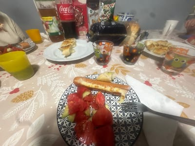 The image shows a table set for a meal, likely at home in Pirdop, Bulgaria. The table is covered with a light-colored tablecloth featuring a pattern of leaves and pinecones. On the table are plates with food, drinks, and cutlery.

In the foreground, a plate contains a sausage topped with melted cheese, served with a salad of chopped tomatoes and cucumbers. Next to this plate is a fork resting on a white napkin.

Further back on the table, there are slices of bread on a white plate. Several glasses are visible, some containing dark liquid. Two of these glasses have a decorative pattern of owls. Also on the table are bottles of Coca-Cola and another drink, a box of cereal, and a bag of snacks.

The presence of these items suggests a casual meal or snack time. The overall scene is domestic and informal.