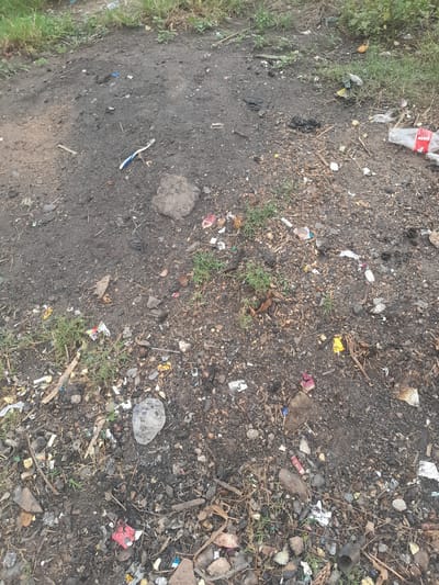 The scene depicts a heavily littered, unpaved ground surface located in Ifo, Nigeria. The ground consists of dark, granular soil interspersed with small stones, pebbles, and sparse patches of green grass. Numerous discarded items are scattered across the area. Visible objects include a partially crushed clear plastic bottle with a red label fragment on the right side, a white and blue plastic toothbrush, multiple fragments of plastic bags and wrappers, torn pieces of colorful paper, and various unidentifiable small debris. Additionally, dried leaves, twigs, and small plant matter are mixed with the soil and litter. A dark, irregular rock or compacted dirt clump is present near the center-left. No individuals or specific actions are depicted.