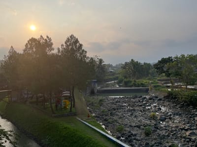 The image captures an outdoor scene at a location in Krasak, Indonesia, during what appears to be early morning or late afternoon due to the low angle of the sun. The sun is visible in the upper left quadrant, casting a warm, soft light over the landscape. In the foreground to the left, a canal or river flows, with a grassy embankment on its right side. This embankment features a small playground with a yellow slide and a red and blue climbing structure. Pine trees and other vegetation line the embankment.
Further into the scene, a concrete dam or structure is visible across a body of water, which appears to be a reservoir behind the dam. The water is a greenish hue. To the right of the dam, the terrain transitions to a rocky, dry riverbed with scattered water puddles. Beyond the dam and riverbed, the landscape opens up to show lush greenery, including palm trees and other tropical foliage, as well as what seem to be agricultural terraces or rice paddies in the distance. Several small structures and buildings are interspersed within the vegetation, suggesting a rural or semi-rural settlement. The sky is partly cloudy, with a hazy, overcast quality. A single bird is visible in flight in the upper right quadrant of the sky. The overall impression is a serene natural landscape with elements of human development, such as the playground and dam.