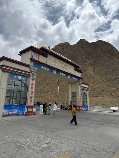 The image depicts an entrance archway to the Qomolangma National Park, with a mountain backdrop. Written on the archway in Chinese are "Zhū zhēn lǎng mǎ fēng guó jiā gōng yuán," which translates to Qomolangma National Park, and "Zhōng huá mín zú yī jiā qīn" meaning "Chinese people are one family."  

Near the arch, there are several people, including a person in a yellow jacket and others wearing white outfits. The park's building, attached to the arch, has a sign reading "Zhōng huá mín zú yī jiā qīn" again.

There is a flag pole extending from the top of the building.
The sky is cloudy.
