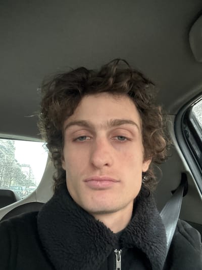 The image displays a head-and-shoulders portrait of a young adult male seated inside a vehicle in Saint-Mandé, France. The subject has medium-length brown, curly hair, light-colored eyes, and a neutral expression, looking directly forward. He is wearing a black jacket with a textured, possibly faux fur or sherpa, black collar and a visible silver zipper pull at the front. A dark seatbelt is secured across his right shoulder and chest. A small, dark earring is visible on his left earlobe. The vehicle's interior is dark, with a grey headliner visible in the upper frame. Through the rear window, an out-of-focus urban background is discernible, showing bare tree branches and light-colored buildings, characteristic of city architecture. Water droplets are visible on the exterior surface of the window, indicating precipitation or condensation. The overall lighting suggests an overcast or low-light exterior condition.