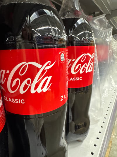 The image shows two bottles of Coca-Cola Classic, packaged together. The bottles are made of dark plastic and are partially filled with a dark liquid. Each bottle has a red label with the white Coca-Cola logo. The label also includes the text "CLASSIC". One of the labels also has nutritional information, showing "105 Kkal" per 250 ml and "0% TOTAL". The bottles are lying horizontally, possibly on a shelf in a store or in a delivery box, given the context of Новомарьевская, Russia. The packaging is clear plastic wrap, also visible. The lighting suggests an indoor environment. No people are visible in the image.