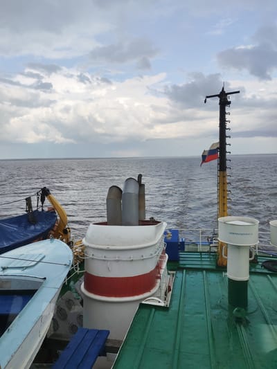 The image depicts a view from the deck of a vessel at sea, likely on a river or large lake, under an overcast sky. The water is choppy. In the foreground is the ship's equipment including its deck painted with dark green with some blue objects. A smokestack painted with red and white stripes sits in the center.  A tall mast displays a Russian flag. The inscription "АККУМУЛЯТОРНАЯ" is visible on a piece of the vessel's structure, which translates to "Battery Room". The location is unlikely to be Veregonets as it is not on a large body of water.