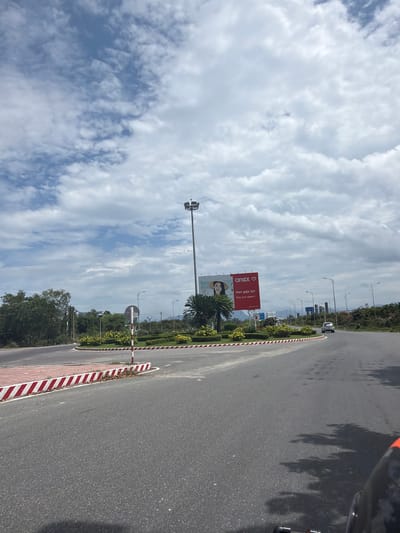 This media file presents a bright, daytime scene of a highway or major road in Cam Lâm, Vietnam, viewed from a slightly elevated perspective, possibly from a vehicle. The road, made of asphalt, extends from the foreground into the distance on the right side of the frame. A median strip, bordered by a distinctive red and white striped curb, separates the road, and features planted bushes, small trees, and palm trees.

The sky, occupying the upper left and central portions of the image, is partly cloudy with large, wispy white clouds against a blue background, suggesting fair weather. Strong shadows of trees and what appears to be a motorcycle or scooter are cast onto the asphalt in the foreground, indicating a sunny day, likely mid-morning or late afternoon.

A prominent red billboard stands on a tall pole on the left side of the median strip, featuring an advertisement. Visible text on the billboard includes the brand name "anex" (accompanied by a heart symbol), the Vietnamese phrase "Hẹn gặp lại!", and its English translation "See you again!". A website "www.anex.com.vn" and a phone number "1900 633 341" are also partly visible. Below the red section, another part of the billboard shows the face of a smiling woman. Further down the road, distant hills or mountains can be seen on the horizon. Streetlights are scattered along the road. The general infrastructure and tropical vegetation are consistent with the provided location context of Cam Lâm, Vietnam.
