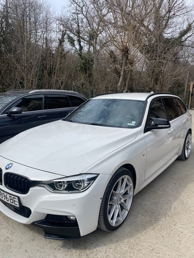A white BMW 3 Series F31 Touring wagon, equipped with a black front grille, LED "angel eye" headlights, a black lower front splitter, roof rails, black side mirrors, and custom multi-spoke silver alloy wheels, is parked on a light gray paved surface. The vehicle displays Bulgarian license plate "T 896 BE" on its front bumper. Partially visible behind and to the left of the BMW is a dark-colored wagon or SUV, also with roof rails. The background consists of a dense arrangement of deciduous trees, mostly bare of leaves, indicating a cool season. The sky is overcast. No individuals are present in the scene, and no discernible actions or interactions are occurring. The location is Kolena, Bulgaria.