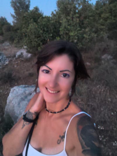 An adult female subject is depicted in a close-up, likely selfie, outdoors in Shaalvim, Israel. She has dark, shoulder-length hair and is smiling, revealing her teeth. Her eyes are adorned with prominent purple eyeliner, and a small fleck of glitter is visible below the outer corner of her right eye (viewer's left). She wears a white spaghetti-strap top, a black beaded choker necklace, and a black band on her left wrist. Dark tattoos are visible on her left upper arm and forearm, and a smaller dark tattoo, possibly the letter 'A' or a similar symbol, is on her upper chest near her right shoulder. Her right hand is positioned against the side of her neck. The background comprises a natural, undeveloped landscape featuring dense green-brown foliage, dry earth, and scattered light-colored rocks. A pale blue sky is visible in the upper left. The lighting suggests dusk or an overcast day.