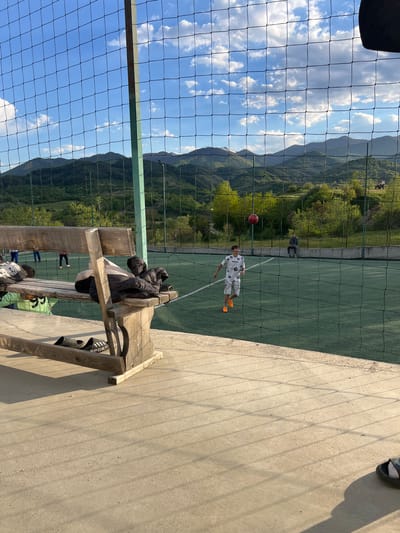 Captured from behind a chain-link fence, the scene presents a verdant valley landscape with visible mountain ranges under a sky dotted with white clouds. A soccer field is enclosed within the fencing, where a young individual wearing a white patterned soccer jersey and orange cleats is captured in action, seemingly heading a red soccer ball. Another person is visible further away on the field, appearing to be sitting or standing. The foreground shows a wooden bench with scattered objects like shoes and a jacket. The location is Kornitsa, Bulgaria.
