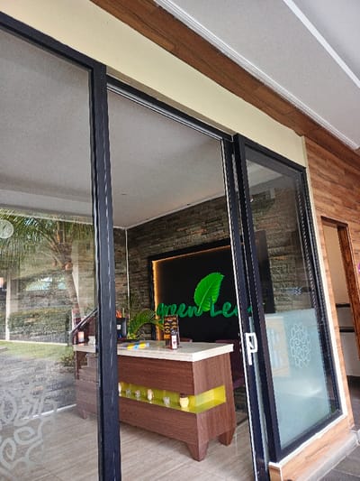 A partially open sliding glass door reveals the interior of a business called "Green Leaf". Inside, a reception desk with a light-colored countertop and brown wood paneling is visible. A black wall behind the desk features a glowing green logo and lettering. Outside, a lush green garden with palm trees can be seen through the glass. The exterior architecture includes wooden-style paneling and a light-colored roof overhang.
