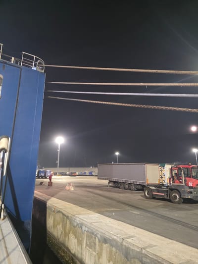 An aerial view of a port at night in Ravenna, Italy, shows large ropes from a ship extending across the frame. In the foreground, a blue ship hull is visible on the left. Further back, a long, grey trailer truck with a red cab is parked on the tarmac. The ground is a dark asphalt surface with tire marks. Numerous large white shipping containers are stacked in the background, along with industrial lighting towers that illuminate the scene. The overall impression is one of a busy, working port operating under the cover of darkness.