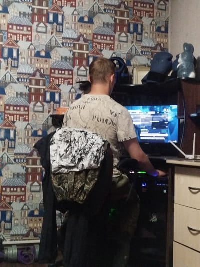 The image shows a person sitting in front of a computer screen, playing a video game. The person is wearing a white t-shirt and is seated in a black chair. The background is a wallpaper with a pattern of houses. There are several objects on a shelf behind the person, including a blue hat and two decorative figures. The computer screen displays a game interface. The scene appears to be indoors, possibly in a residential room. The location in Kirensk, Russia, cannot be determined from the visual information provided in the image.