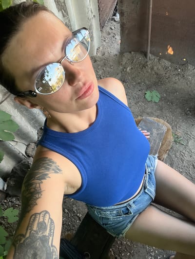 A woman is lying down outdoors, with a close-up shot of her upper body. She is wearing a blue tank top and denim shorts. Her face is turned towards the camera, and she is wearing large, round sunglasses that reflect the surroundings. Her lips are slightly parted. She has tattoos on her left arm. The setting appears to be rustic and outdoors, with dirt, leaves, and a wooden surface visible. A metal object is also present in the background. The lighting suggests it is daytime.