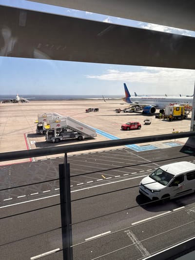 The image depicts an active airport apron at Tenerife South Airport (TFS) in Granadilla de Abona, Spain, viewed from an elevated position through a large window. The foreground features a dark metal railing with multiple horizontal bars.
In the mid-ground, a white "Jet2.com" and "Jet2holidays" branded aircraft, likely a Boeing 737, is parked on the tarmac with its blue and orange tail section oriented towards the right. A yellow ground power unit (GPU) or similar utility vehicle is positioned near the aircraft's starboard side, with at least one individual, appearing to be an airport worker, standing nearby. Several baggage carts and ground handling equipment are also located near this aircraft. A red "Jet2holidays" branded service car is parked on the apron in front of the aircraft. Two mobile passenger stairs, white with "AP" branding, are parked on the tarmac to the left of the main aircraft.
Further in the distance, a second aircraft with its nose facing the viewer is visible near a taxiway or runway, close to the ocean. The ocean forms a distinct horizon line under a clear blue sky with minimal cloud cover, indicating sunny daytime conditions. The tarmac is marked with white and blue lines, delineating vehicle paths and aircraft stands.
In the immediate foreground, below the metal railing, a white panel van, also bearing "AP" branding, is visible on a paved service road. The van is partially obscured by the railing structure. Reflections from inside the viewing area are visible on the window glass, particularly in the upper portion of the image.