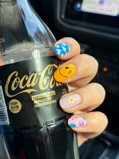 A close-up shot shows a hand holding a plastic bottle of Coca-Cola Zero. The focus is on the hand and the bottle, revealing several details. The hand's fingernails are painted in various playful designs: one features blue with white clouds, another a yellow smiley face, a pale pink nail with a white flower, and a pink nail adorned with small pink and blue smiley faces.
The Coca-Cola bottle is dark with gold lettering, indicating "Coca-Cola zero caffeine zero sugar zero calories." A label on the bottle states "new look SAME TASTE." A barcode is also visible at the bottom of the label.
The background is dark and indistinct, except for a small area showing a blurred screen, likely of a navigation device, suggesting the scene is set inside a vehicle.
Based on the OCR result there is no city of Berģi, Latvia visible in the image.