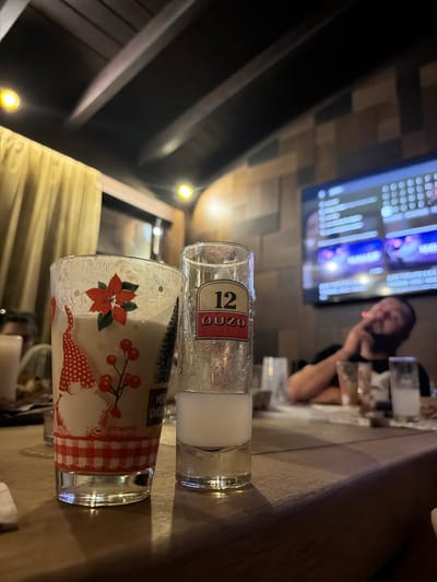 The image depicts an indoor scene within a commercial establishment, specifically a restaurant or bar, located in Petrich, Bulgaria. In the foreground, positioned on a polished wooden table, are two drinking glasses. The glass on the left is a festive tumbler with a white, frosted upper portion and a clear base. It is adorned with a red poinsettia, a gnome in a red conical hat, red berries on a branch, a small pine tree, and a red and white gingham pattern along its base, with the partial word "CERVE" visible. This glass contains an opaque, light-colored liquid. To its right, a clear shot glass branded "12 OUZO" in red text on a white label holds a milky white, opaque liquid and exhibits condensation.

In the mid-ground, out of sharp focus, a male individual with a beard is seated at the table, leaning back slightly. He is holding an object to his mouth, from which vapor or smoke is visible, suggesting he is smoking or vaping. He is looking towards a large, rectangular flat-screen television mounted on the wall behind him. The television screen displays blurred electronic content, possibly a menu or video. The wall features a textured pattern of various rectangular panels. The ceiling is dark with visible structural beams and is illuminated by several bright, circular yellow light fixtures. A light-colored curtain is visible on the left side of the frame. Additional blurred glassware and table settings are present on the table in the background.