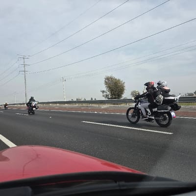 A highway scene under a muted sky. The viewpoint appears from inside a car, indicated by the blurred red dashboard. Several motorcycles are traveling along the road. One prominent motorcycle carries two helmeted riders, equipped with luggage racks and bags. The remaining motorcycles are farther ahead. Overhead, a grid of power lines stretches across the sky. The roadside features a safety barrier and some sparse vegetation.