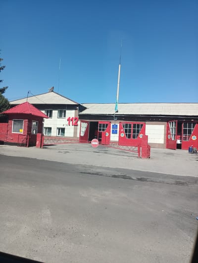 The image depicts the exterior of a fire station located in the city of Talgar, Kazakhstan, under a clear blue sky. In the foreground, a paved road leads towards the building. To the left, a small red guardhouse with a glass window is visible, and in front of it, a red brick wall. A red gate separates the road from the fire station complex.
The fire station itself is a long, light-colored building with a corrugated roof. Large red garage doors are a prominent feature, with three doors clearly visible. Two of these doors are open, revealing glimpses of the interior. The number "112" in large red numerals is displayed on the front of the building. The numbers "2" and "3" are also visible on the garage doors. A flagpole with a flag is present on the roof, along with some antennas. A red sign with a white circle and a diagonal line, indicating "no entry," is placed on the right side of the gate. Various objects, including a blue bucket and a red container, are scattered near the open garage doors. A satellite dish is visible on the roof of the main building. The overall scene suggests a functional fire station in a dry, sunny environment.