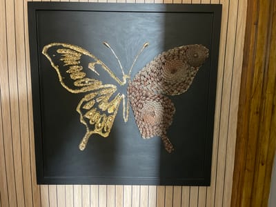 A framed artwork of a butterfly is displayed on a wall covered in vertical wood paneling. The butterfly artwork has a black background. The left side of the butterfly, representing its wings, is adorned with gold glitter and appears to be made of a textured material. The right side of the butterfly, also representing its wings, is constructed from what appear to be pinecone scales, arranged in a spiral pattern. The butterfly's body is also made of pinecone scales, and its antennae are thin and metallic gold. The wood paneling on the wall is light in color with dark vertical lines, and to the right of this, there is another section of wood paneling that is darker and more golden-brown in color. This artwork is located in the city of Portsmouth, United Kingdom.