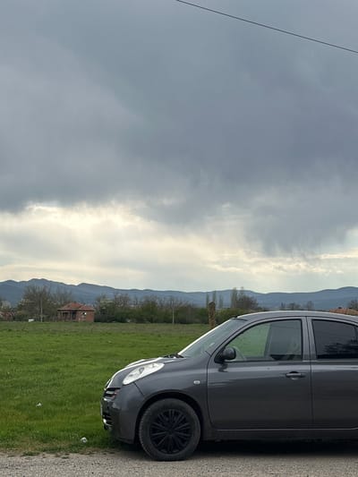 This media file presents a rural or semi-rural landscape in Padarevo, Bulgaria, captured on an overcast day. The primary subject is a dark grey hatchback car, likely a Toyota Yaris, parked on a gravel road or dirt path that occupies the right side of the frame. The car is viewed from its front-right side, featuring dark tinted windows and black multi-spoke wheel rims.
To the left of the car, a lush green grassy field extends into the midground, indicating springtime. Scattered trees, some with emerging leaves and others still bare, dot the landscape. In the distance, a small, old brick house with a reddish-orange tiled roof is visible, partially obscured by foliage and situated against a backdrop of undulating hills or mountains. The mountains stretch across the horizon under a cloudy sky.
The sky is largely covered by grey clouds, with some lighter patches suggesting diffused sunlight. A single black power line or cable runs diagonally across the upper left portion of the image. No people are visible, and the car appears to be stationary. A few pieces of litter are discernible on the ground near the car. No text is visible in the image. The scene conveys a quiet, natural setting with human elements integrated into the landscape.