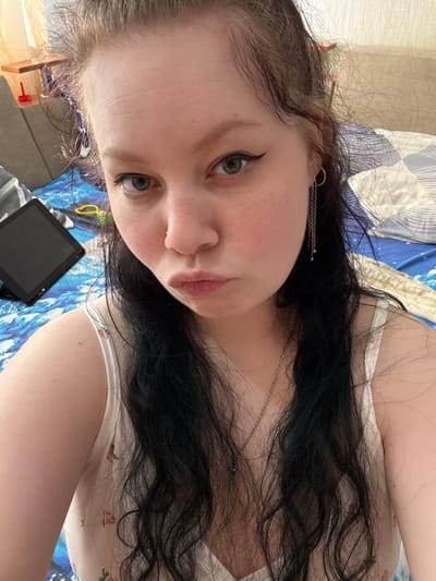 A young woman with dark, wavy hair is looking at the camera and puckering her lips. She is wearing a white tank top with a floral pattern and a silver necklace. She has on eyeliner and a small silver hoop earring with a chain. A tablet is visible on a blue and white patterned surface behind her. The scene appears to be indoors, possibly a bedroom. The image was taken in the village of Vylygort, Russia.