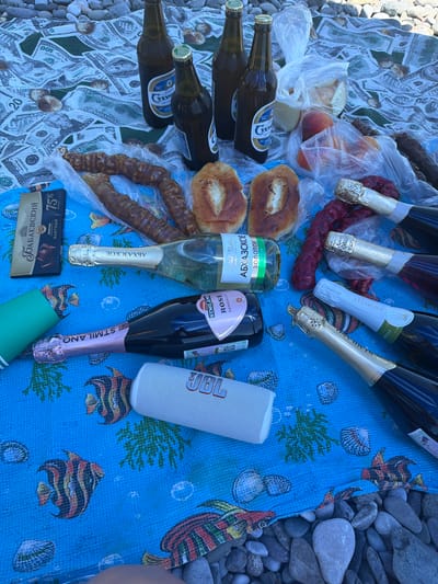 This overhead shot captures a lively picnic spread on a pebble beach, likely in Amzara, Abkhazia. The scene is dominated by a collection of beverages, including several bottles of what appears to be sparkling wine or champagne, some in gold and silver foil, and a few dark glass bottles that might contain beer. Alongside the drinks are various food items: plump peaches in plastic packaging, two golden-brown pastries with visible white filling, and two long, twisted sausages. A bar of chocolate with "75% kakao" and "Rabaevsky" in Cyrillic lettering is also present.
The picnic blanket is a vibrant blue with a whimsical pattern of colorful fish, shells, and seaweed, contrasting with the natural grey and white pebbles of the beach that surround it. A white cylindrical speaker, with "JBL" visible on it, sits on the blanket, suggesting music is part of the gathering. A few bottles of dark liquid, likely beer, are placed on a background that resembles US dollar bills, adding an unusual element to the scene.
While no people are directly visible, the arrangement of food and drinks implies a shared meal or gathering. The bright sunlight suggests it's daytime, and the setting on a beach with numerous pebbles indicates a coastal or lakeside location. The overall mood seems relaxed and celebratory, a classic picnic enjoyed outdoors. The visible text, primarily on the beverage labels and the chocolate bar, is mostly in Cyrillic, reinforcing a potential location in a Russian-speaking or Eastern European region.