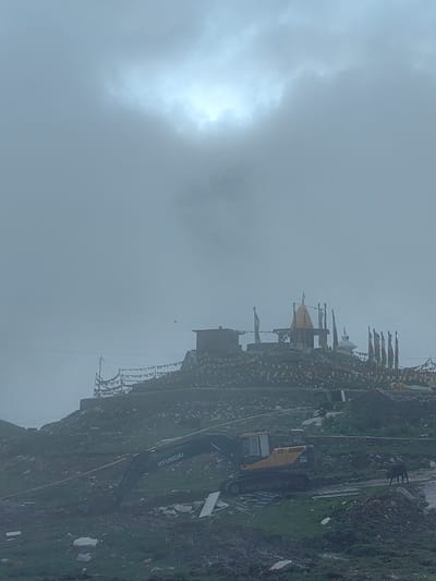In Marhi, India, a temple complex stands atop a hill, adorned with prayer flags. Below, a yellow Hyundai excavator is positioned on a muddy, construction site. The sky is overcast with the sun partially obscured by clouds. A dark-colored animal, possibly a yak or cow, is visible in the distance to the right of the excavator. The terrain is rocky and uneven, with some debris scattered around the construction area.