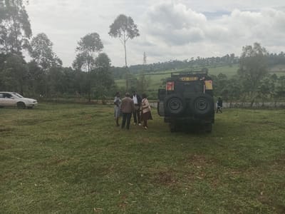 The image shows a group of four individuals gathered near a dark-colored, possibly military-style, vehicle in a grassy field. The vehicle has two spare tires mounted on the rear and a license plate reading "GKB 800J". To the left, a silver sedan is parked on the grass.
The group of people consists of three women and one man. They appear to be engaged in conversation or discussion. One woman is wearing a patterned dress, another is in a brown skirt and light top, and the man is wearing a suit jacket.
The location is Moticho ward, Kenya, suggested by the landscape which includes rolling hills, trees, and a partly cloudy sky. The background includes vegetation and what may be a distant treeline. The image appears to be taken during daylight hours under an overcast sky.