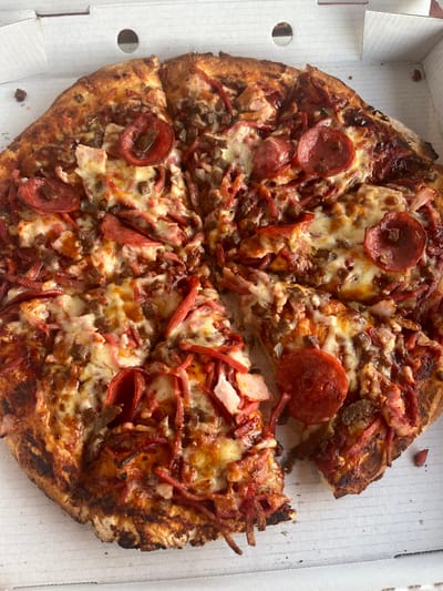 An overhead view depicts a large, round, cooked pizza contained within a white cardboard pizza box in Applecross, Australia. The pizza is generously topped with melted, golden-brown cheese, multiple round slices of pepperoni, crumbled ground meat (likely beef or sausage), and shredded or diced pieces of red meat (possibly ham or bacon). The red sauce base is visible beneath the toppings. The pizza crust is thick, golden-brown, and exhibits some darker, charred areas around its periphery. The pizza has been cut into eight slices, with one slice removed from the lower right quadrant, leaving a void. Small food debris is present on the white surface of the pizza box. No individuals are visible, and no actions are currently taking place in the static scene.