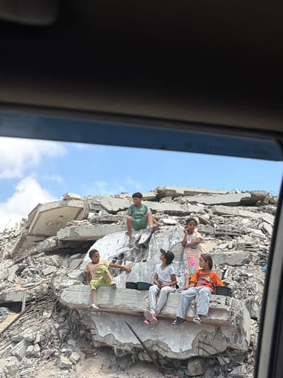 In Jabalia, Palestinian Territories, a group of children sits amidst the rubble of destroyed buildings. The scene is one of devastation, with concrete debris, twisted metal, and exposed rebar dominating the landscape. The sky above is a clear blue, providing a stark contrast to the destruction below.
One boy, wearing a green t-shirt with the word "REFLEX" on it, sits on a large concrete slab, his legs dangling. To his right, a younger boy in a yellow t-shirt and patterned pants perches on a different piece of debris, looking towards the side. In the middle ground, a girl in a light-colored t-shirt and gray pants sits with a black baseball cap on her head, looking upwards. Next to her, another girl in a light pink t-shirt with colorful graphics and white pants stands with her hands clasped. To her right, a girl in an orange t-shirt sits with a black bucket at her feet.
The children appear to be interacting, with some looking at each other and others looking outwards. The overall impression is one of resilience and adaptation in the face of severe destruction, with the children finding a place to rest and gather amidst the ruins. The image was captured from within a vehicle, as evidenced by the dark frame and tinted glass.