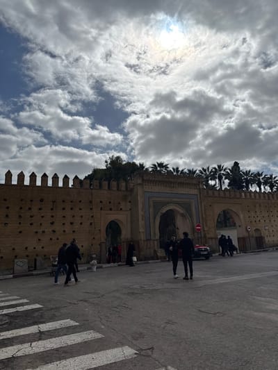 This image depicts a daytime scene in Mechouar Fes Jdid, Morocco, featuring an ancient fortified wall and ornate gateway under a partly cloudy sky. The sky is dominated by large, scattered white and grey cumulus clouds, with the diffused light of the sun visible through a cloud in the upper-center.
The prominent feature is a long, light-brown historical wall stretching across the midground. Its top edge is adorned with crenellated battlements. The wall's surface is marked with numerous small, dark square indentations. A smaller, plain arched entry is located within the wall on the left side. Centrally positioned is a larger, ornate arched gateway featuring intricate geometric tilework in blue, white, and brown hues. To the right of this main gate, a white circular road sign with a red border indicating "No Entry" is visible. Beyond the wall and gates, a line of tall palm trees and other green foliage is visible against the sky.
The foreground consists of a dark grey asphalt road, exhibiting signs of wear. White, cracked pedestrian crossing stripes are visible in the lower-left corner. Approximately 15-20 individuals are present in the scene. On the left side of the road, three men are moving to the right; one is pushing a two-wheeled handcart, and a white bucket is on the adjacent sidewalk. A small white heart outline is spray-painted on the lower part of the wall. In the center-right, two men in dark clothing stand facing each other on the asphalt, appearing to be engaged in conversation. A dark-colored sedan is partially visible behind them. Further right, a group of 5-7 individuals is congregated near another opening in the wall. Individuals are generally observed walking, standing, or interacting in small groups.