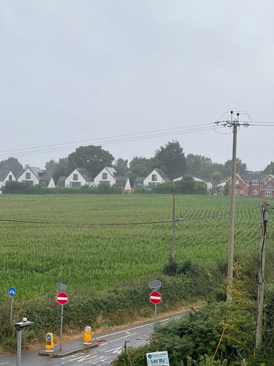 The image displays an overcast rural-urban fringe landscape in the Tithebarn area of Exeter, United Kingdom. In the foreground, a wet asphalt road or path runs horizontally, bordered by green foliage. On the left side of the path, a paved section features a blue circular sign with a white bicycle symbol, two yellow bollards, and a street light pole. A red circular 'No Entry' sign is centrally positioned on this paved area. Further right, another red circular 'No Entry' sign with a solar panel mounted above it stands between the path and the field. A "Let By" sign for "Estuary Lettings" is partially visible in the bottom right corner.
The midground is dominated by a large, cultivated field of dense green crops, likely corn or maize, arranged in visible rows receding towards the background. Overhead power lines traverse the upper portion of the image, supported by two wooden utility poles; a thicker pole with electrical equipment is on the right, and a thinner pole is further left within the field. In the background, a line of mature trees partially obscures and frames a residential area. Several modern, white-rendered houses with dark, steep gabled roofs and prominent dark-framed windows are visible behind the field. To the far right, a traditional red-brick house with a dark roof is also present. The sky is uniformly light grey and overcast. No individuals, animals, or vehicles are present in the scene.