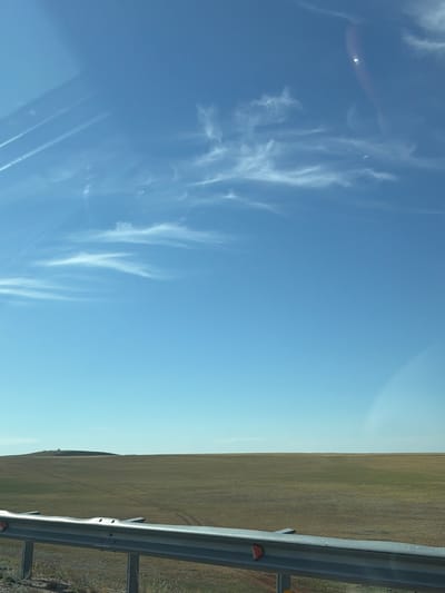 This is a wide-angle, eye-level photograph taken from a vehicle, looking out over a vast, flat landscape. The top two-thirds of the image is dominated by a clear blue sky with wispy white clouds and what appear to be contrails. In the lower third, a wide, flat field of dry, light brown and green vegetation stretches to the horizon. A metal guardrail, likely from a road, runs along the bottom of the frame. The landscape suggests a rural or steppe environment, characteristic of the region around Торғай стансасы, Kazakhstan. There are no people or animals visible in the scene. The lighting indicates it is daytime, likely mid-morning or afternoon.