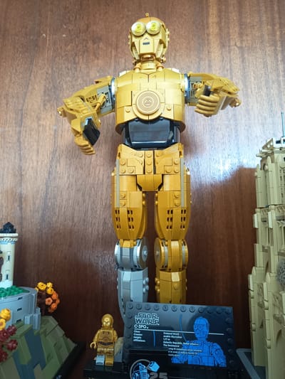This media file showcases a collection of Lego Star Wars models displayed against a wooden background. The central subject is a large, articulated Lego C-3PO figure, gleaming in its signature gold color. To the left, a smaller Lego model depicts a lighthouse-like structure with a green, rocky base adorned with Lego foliage and flowers. To the right of the large C-3PO, a beige Lego building, reminiscent of Tatooine architecture, is partially visible.
In the foreground, a smaller, classic Lego C-3PO minifigure stands on a black display base. This base features a graphic detailing information about the character, with visible text including "STAR WARS", "C-3POTM", "Class: Protocol Droid", "Creator: Anakin Skywalker", "Height: 1.77 m", and "Affiliations: Galactic Republic, Rebel Alliance, The Resistance". Other text on the display is too small to read clearly.
The lighting suggests an indoor setting, possibly a display case or shelf, with the wooden texture of the background providing a warm and rustic context. The overall scene is static, representing a curated display of Lego creations rather than an event in progress. The focus is on the detailed craftsmanship of the Lego models and the iconic Star Wars characters they represent.