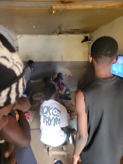 In Bokkos, Nigeria, a group of young individuals are gathered in a room, likely a dwelling or community space, with a low ceiling and unpainted walls. A television is visible, suggesting a shared activity like watching a program or playing a video game. The lighting appears dim, casting shadows and creating a slightly obscured view of the scene. One person in the foreground wears a black t-shirt and has their back to the camera, facing the television. Another person is seen from the back wearing a white t-shirt with dark lettering that reads "BACK TO TRYIN". A few other individuals are seated or standing, their faces obscured by darkness or their orientation. The overall impression is of a casual, informal gathering, typical of community interaction in such an environment.