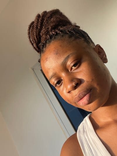 A close-up, slightly angled portrait of a person with dark skin and braided hair. The face is the focal point, revealing a nose ring and an ear piercing. A white tank top peeks into the frame on the right. Sunlight highlights the skin. The background is a combination of a window frame and a white ceiling. The overall composition creates a casual and intimate feel.