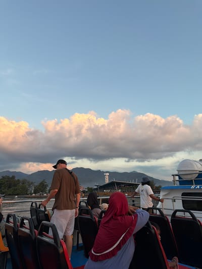 In the city of Pemenang Timur, Indonesia, a group of people are on the deck of a ferry at sunset. The sky is a vibrant blue, with soft pink and orange clouds drifting across the horizon. In the distance, mountains are silhouetted against the colorful sky. The ferry is moving, with the water rippling around it. People are seated on the ferry, facing forward, enjoying the scenic view. A man in a brown t-shirt and grey shorts stands near the front of the ferry, holding onto a railing. A woman in a red hijab is seated, looking out at the scenery. A man in a white t-shirt and a hat stands further back on the ferry, also looking out. The ferry appears to be docking at a port, with buildings and a lighthouse visible on the shore. The overall atmosphere is peaceful and serene, with the warm glow of the setting sun creating a beautiful backdrop for the scene.