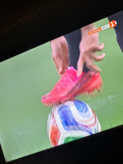 The image shows a close-up of a soccer ball on a green surface, possibly a soccer field. A bright pink soccer shoe is placed on top of the ball, with its cleats digging slightly into the ball's surface. A person's hands are visible in the background, appearing to be in the process of adjusting the shoe or ball. In the upper right corner of the image, there is a broadcast graphic with the text "НА ЖИВО · Б-Н-Т · 1" in orange and white, indicating a live broadcast on BNT (Bulgarian National Television) channel 1. The image is likely a still from a soccer match or training session being televised live. There is no visible indication of the specific location within Miriantsi, Bulgaria.