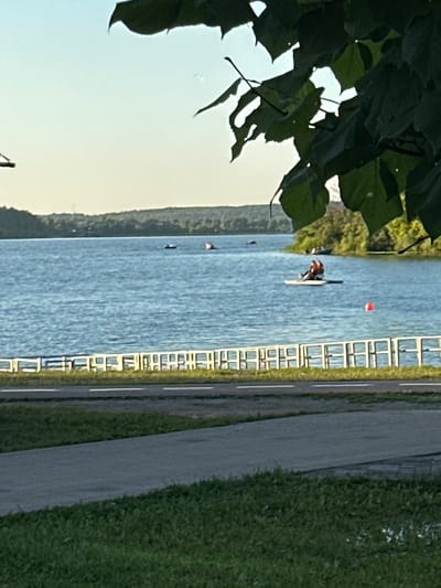 The image depicts a serene waterscape in Solnechnogorsk, Russia, likely during late afternoon or early evening based on the soft light. In the foreground, a paved path runs parallel to a stretch of green grass, suggesting a park or recreational area. Beyond this, a white fence delineates the edge of the path from the water. The vast expanse of a calm blue lake dominates the midground, with several small boats and a person on a paddleboard visible. The paddleboarder is seated and appears to be actively paddling. In the distance, a treelined shore forms the horizon, with a faint hint of buildings or structures. Overhanging the scene from the top of the frame are dark green leaves and branches, partially obscuring the sky and creating a natural framing effect. The sky is a pale, clear blue, transitioning to a lighter hue near the horizon. The overall impression is one of tranquility and outdoor leisure.