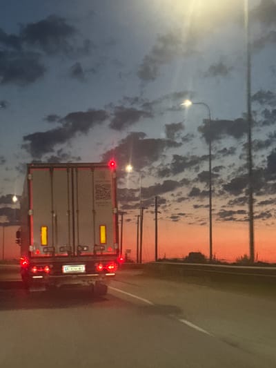 The image captures a rear view of a white semi-trailer truck traveling on a highway at dusk. The truck's brake lights and hazard lights are illuminated, indicating its presence on the road. The sky displays a gradient of colors from a pale blue at the top to a vibrant orange and pink near the horizon, characteristic of a sunset. Scattered dark clouds are visible against the lighter sky. Several tall streetlights line the right side of the highway, with one appearing to be illuminated. The road surface is asphalt with a white line marking the lane. The truck's license plate, partially visible, reads "AA 8744 XX". The scene suggests a journey on a highway, possibly on the outskirts of Korostyshiv, Ukraine, given the context provided.