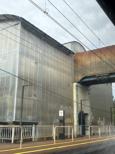 The image shows an exterior view of a building in Pesochny, Russia, taken from what appears to be a moving vehicle, likely a train. The building features a semi-cylindrical structure with a translucent facade, exhibiting signs of wear and staining. An overhead pipe system runs along the top of the building. A platform with metal railings and a yellow safety line borders the building. A small SOS information panel is attached to the building. Overhead power lines are visible against a cloudy sky.