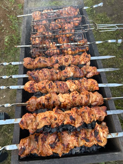 The image shows a close-up, overhead view of shashlik, a type of grilled meat skewer, being cooked in a brazier filled with hot coals. Several skewers are laden with chunks of meat, some of which are slightly charred, indicating they are well-cooked. To the left, some skewers are placed in a grill basket, also filled with meat. Wisps of smoke rise from the coals, suggesting the cooking process is active. The background consists of dry grass and dirt, indicating an outdoor setting, likely in a rural or park area. The lighting suggests it is daytime, possibly late afternoon given the warm tones of the meat and coals. There are no people visible in the frame, but the presence of the grill and the food implies a gathering or a meal preparation. The overall scene conveys a sense of an outdoor barbecue, with the focus entirely on the appetizing appearance of the grilling meat. The location context provided places this scene in Sibay, Russia.