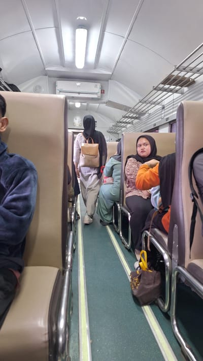 The image depicts the interior of a train car, likely in Karawang, Indonesia, based on the user's prompt. The train car features rows of beige-colored seats with metal framing. The aisle floor is a dark green with yellow and white striping. Overhead, fluorescent lights illuminate the space, and a silver air conditioning unit is mounted on the ceiling.
Several individuals are visible. In the foreground to the left, a person wearing a dark blue hoodie is partially visible from the shoulders down, seated. Further down the aisle, a person wearing a black hijab and a pale grey and white outfit is walking away from the camera, carrying a tan backpack. To the right of the walking individual, a person in a mint green head covering and outfit is seated. In the right foreground, a woman wearing a black hijab and a patterned pink and white shirt is seated, with her hands clasped in her lap. Next to her, another person is seated, wearing an orange long-sleeved shirt and a black head covering. A brown shopping bag with yellow lettering sits on the floor between the seats. The scene appears to be an ordinary journey on a public train.